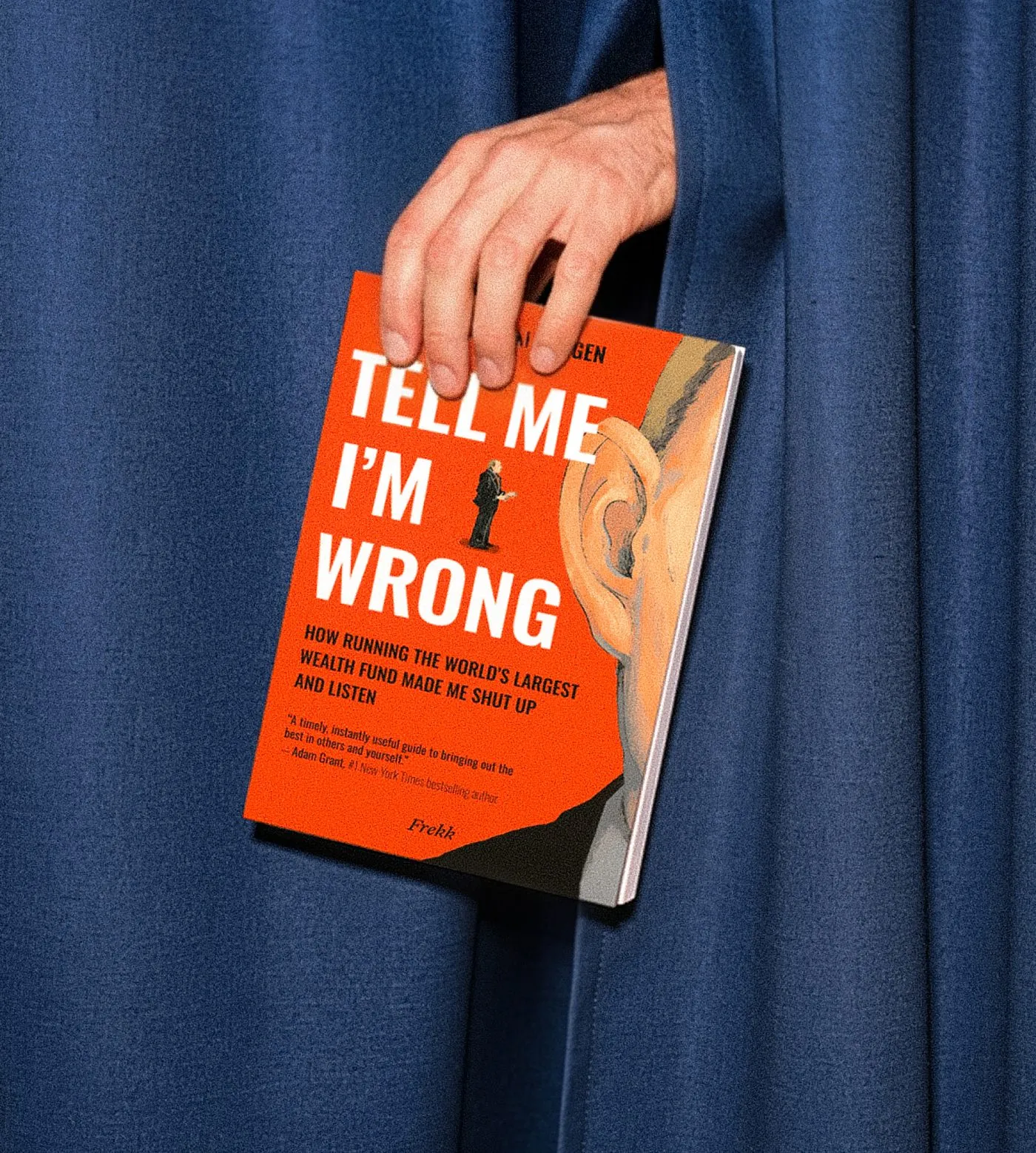 A hand holding a copy of Tell Me I'm Wrong, drawing it out from between blue curtains.